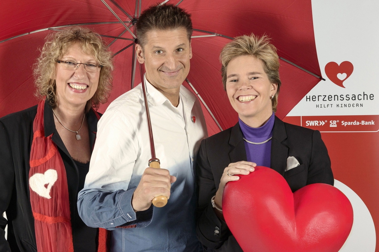Unter einem Schirm (von links): Gitta Haucke (Geschäftsführerin Herzenssache e.V.), Hartmut Engler (Pur) und Dr. Simone Schelberg (Vorsitzende Herzensache e.V. und SWR-Landessendedirektorin Rheinland-Pfalz)
