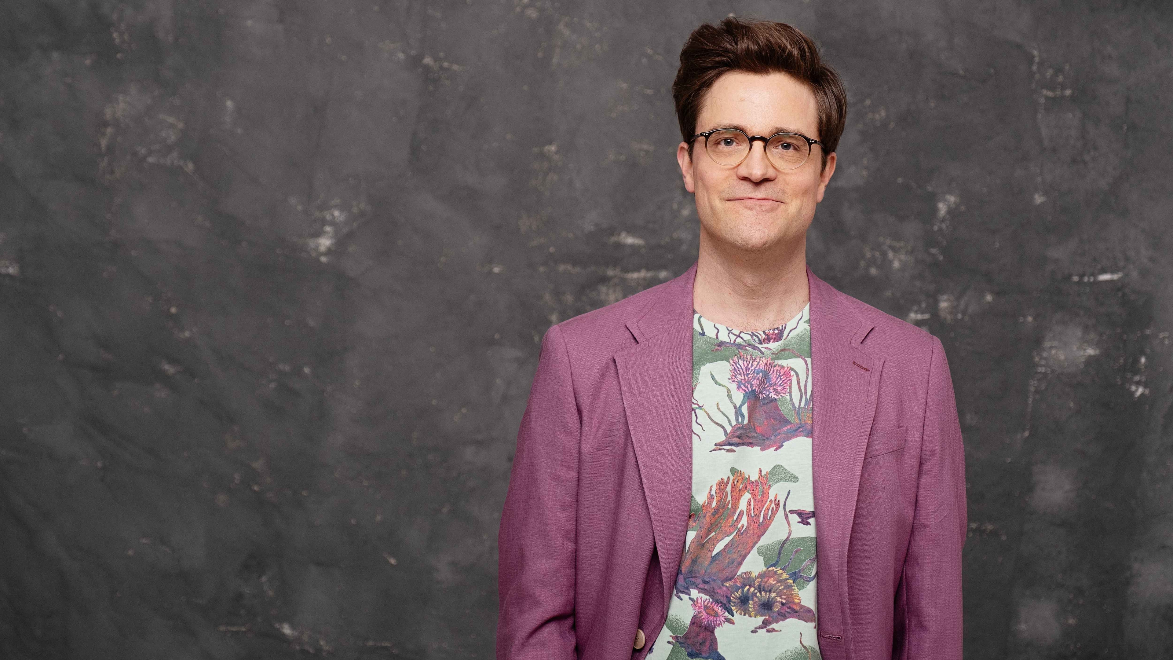 Zeigt Bodo Wartke im rosafarbenen Anzug smart lächelnd vor einer grauen Wand | Abdruck nur gegen Nennung des Fotografen: Sven A. Hagolani | Weitere Informationen unter www.bodowartke.de/presse